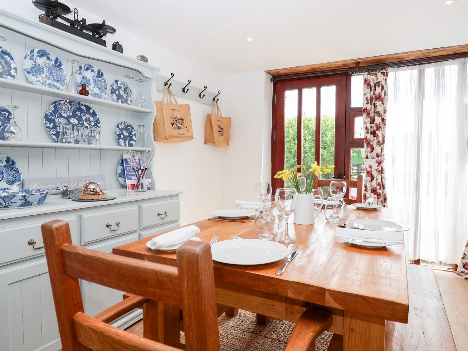 A dining room with a wooden table set with plates glasses and a vase of flowers beside a cabinet with blue and white plates at The Bolthole in Throwleigh near Chagford