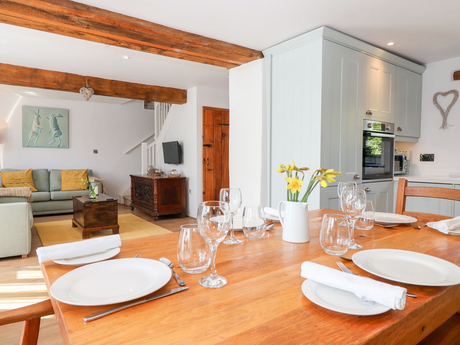 A dining table set with plates glasses and napkins with yellow flowers in a jug in a kitchen living area with wooden beams at The Bolthole in Throwleigh near Chagford