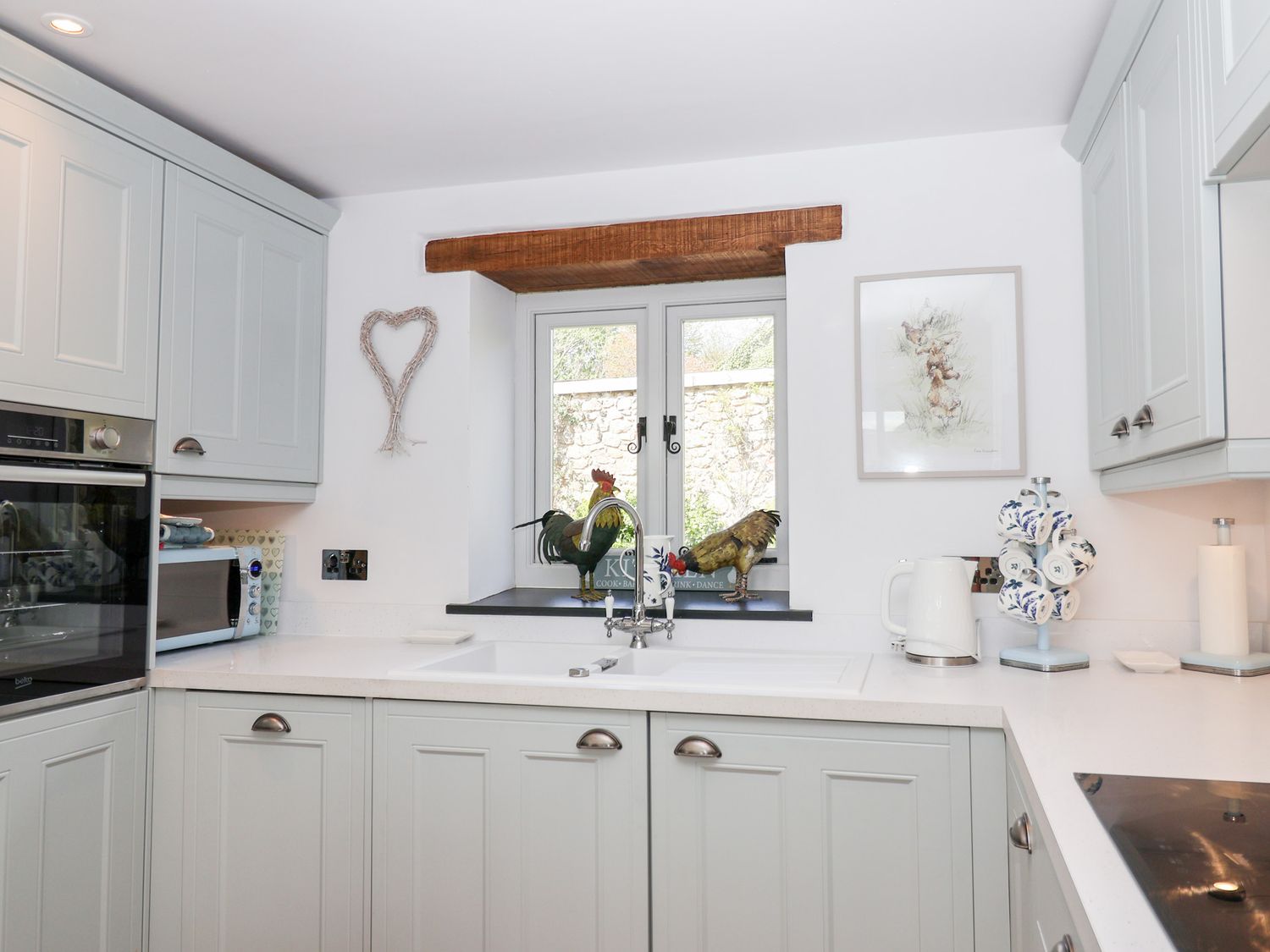 A kitchen with white cabinets a sink under a window two rooster figurines a kettle and a cup holder at The Bolthole in Throwleigh near Chagford