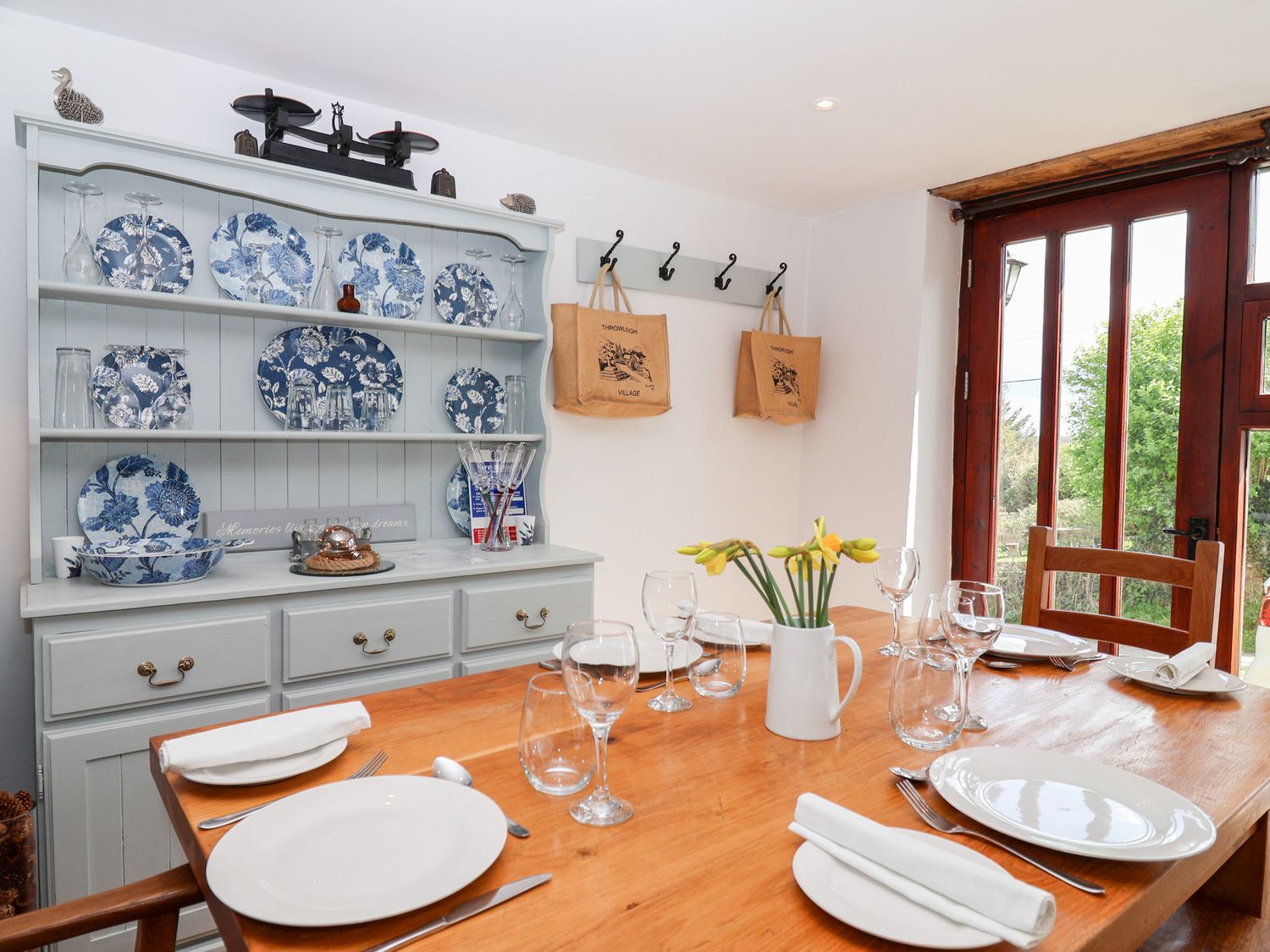 A dining table set with plates glasses and cutlery next to a cabinet with blue and white dishes and a door showing greenery at The Bolthole in Throwleigh near Chagford