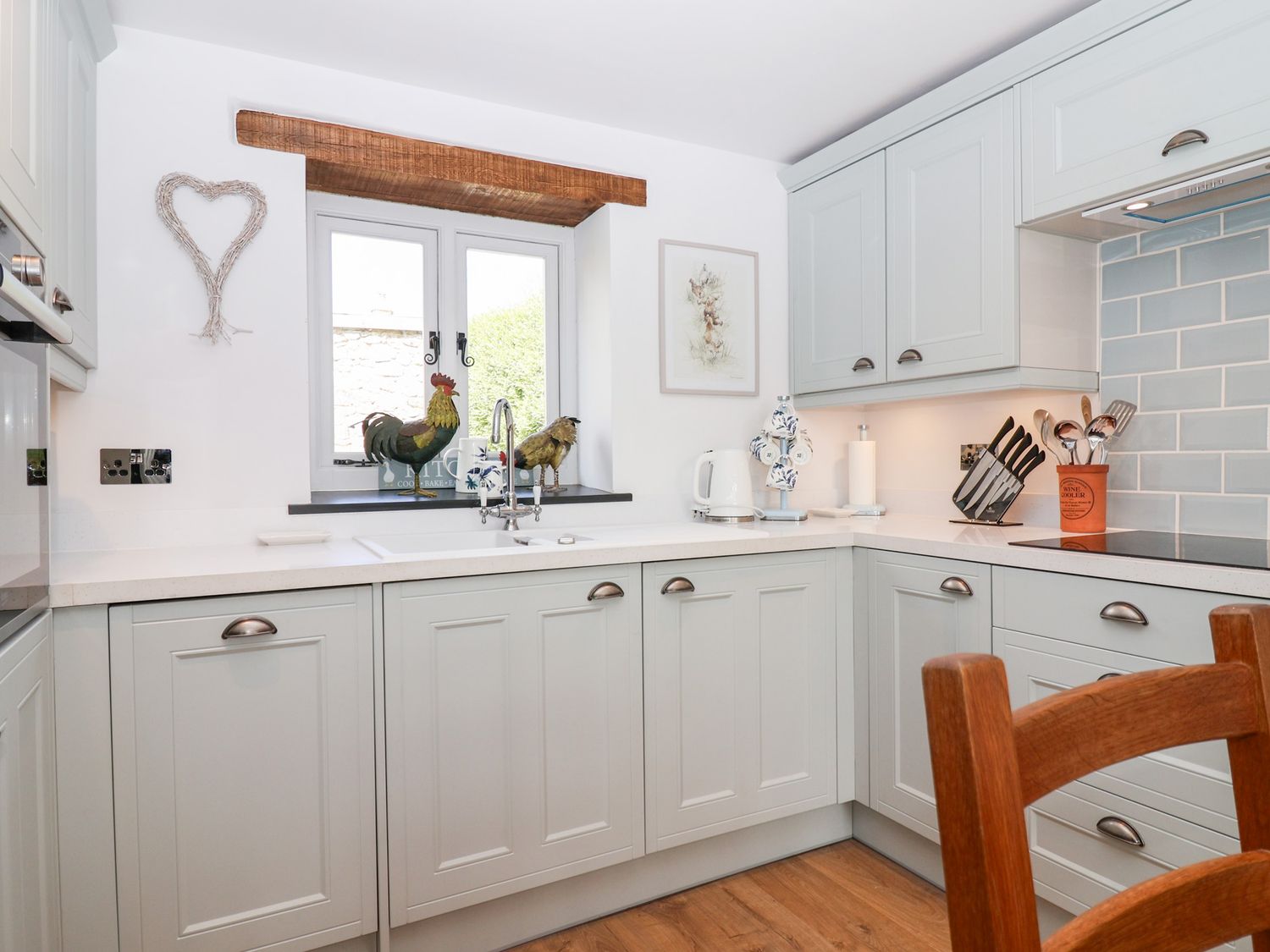 A kitchen with grey cabinets a wooden chair and two rooster figurines on the window sill at The Bolthole in Throwleigh near Chagford