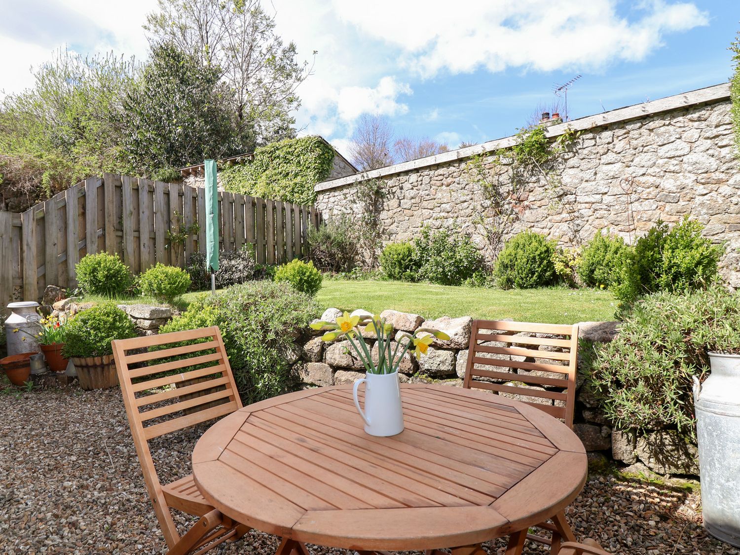 A wooden outdoor table with two chairs and a vase of flowers in a garden with stone walls and greenery at The Bolthole in Throwleigh near Chagford