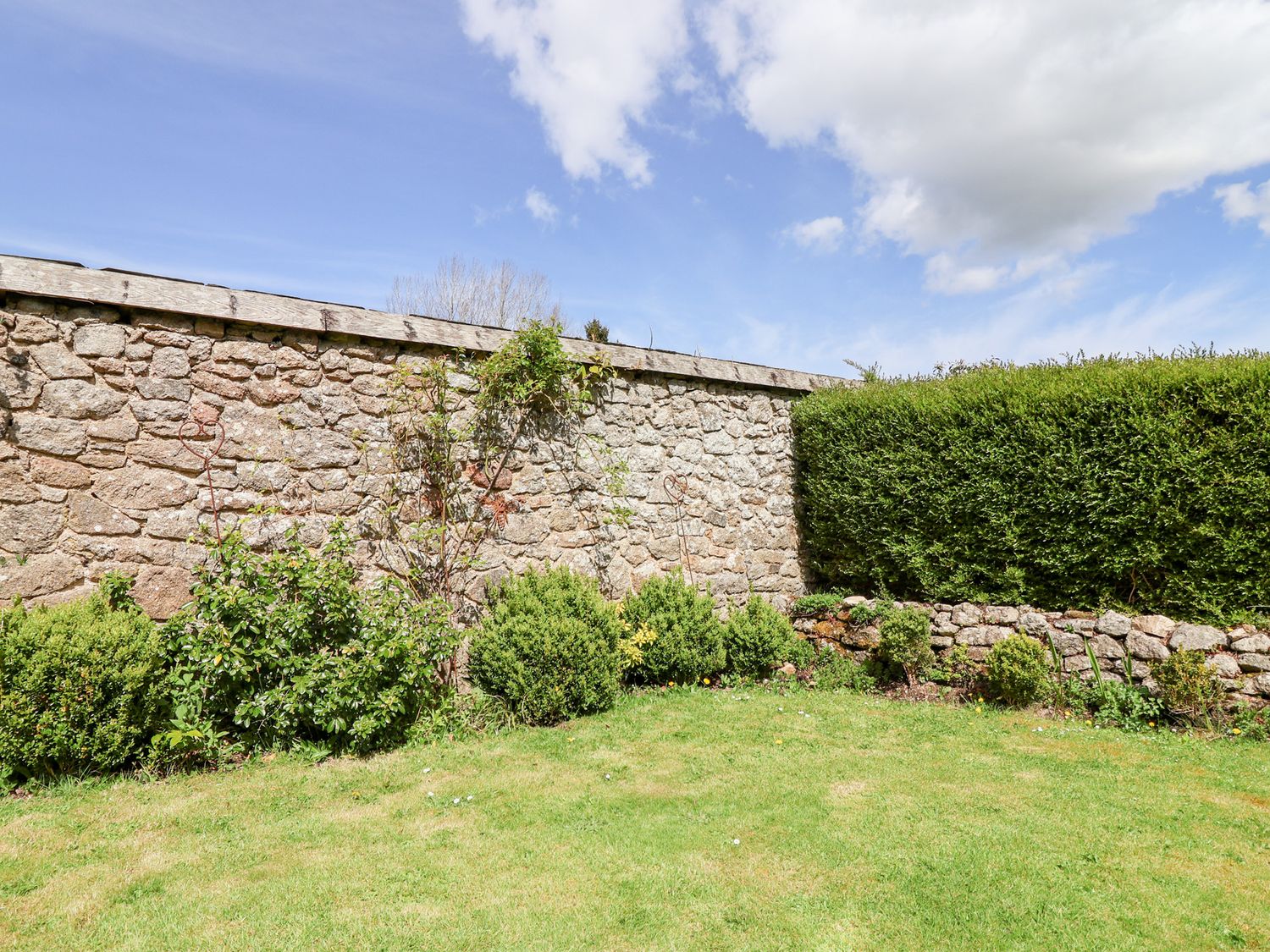 A garden with stone wall shrubs and grass under a partly cloudy sky at The Bolthole in Throwleigh near Chagford