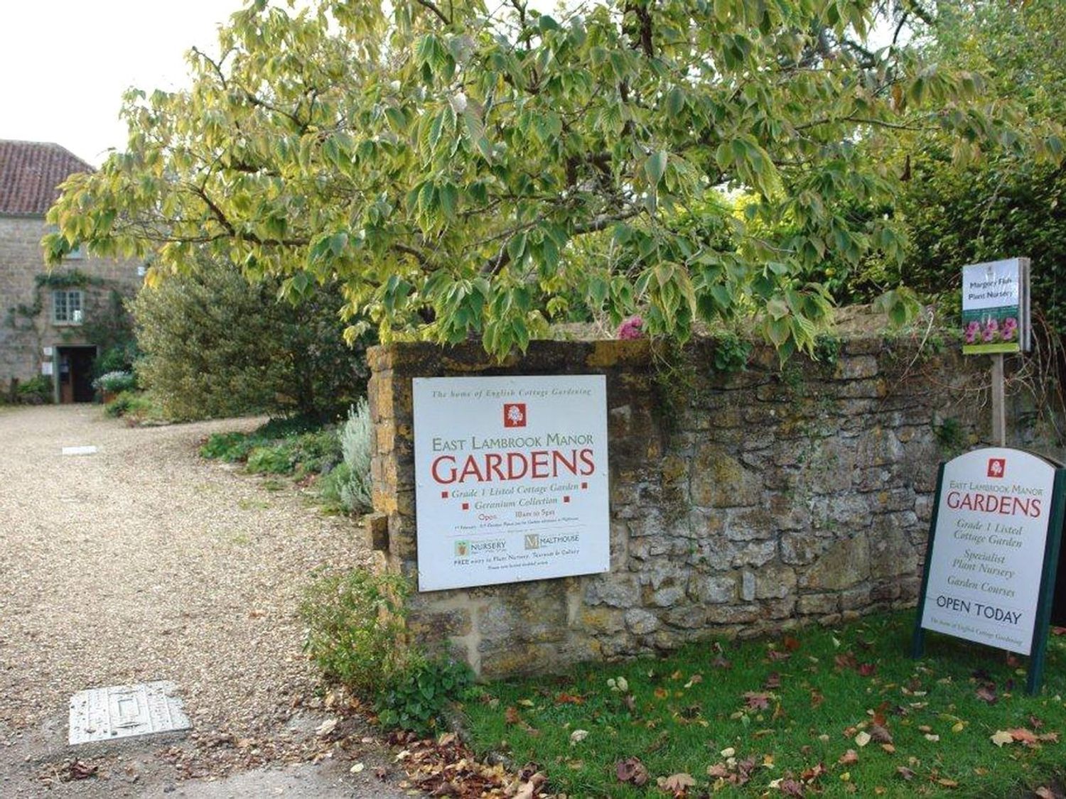 Stone wall and signs for East Lambrook Manor Gardens with a gravel path and plants at East Lambrook Manor Gardens in South Petherton