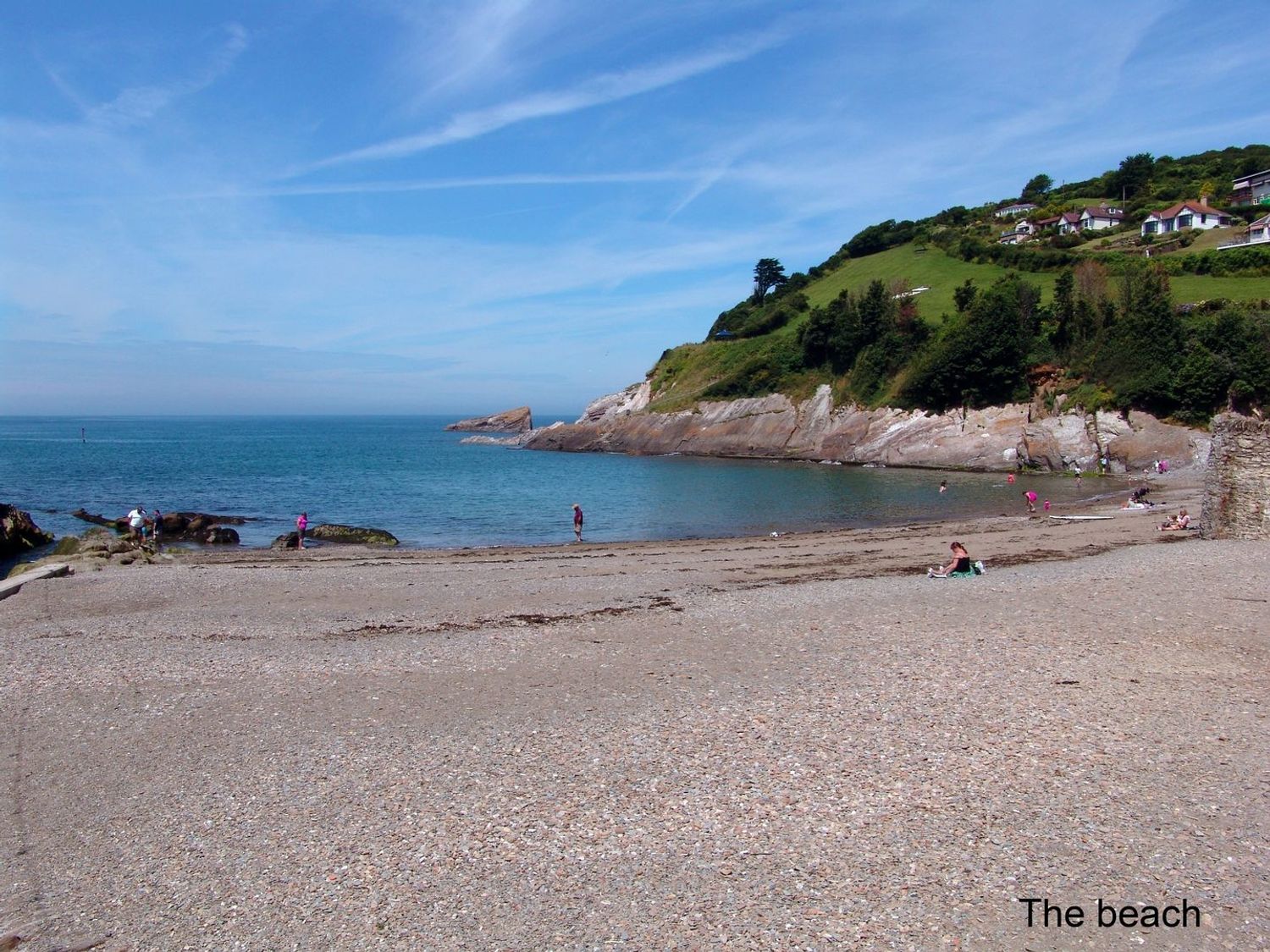 A pebble beach with people near the water and houses on a grassy hill at Owls Roost in Combe Martin