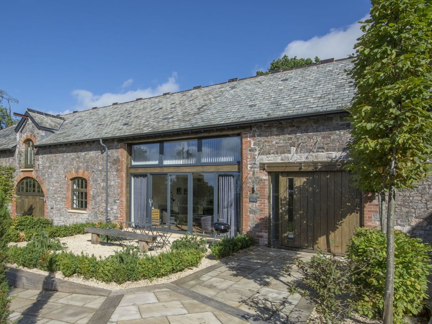A stone and brick house with large glass doors wooden doors and a garden with small plants and a tree at Barley House in Talaton near Ottery St Mary