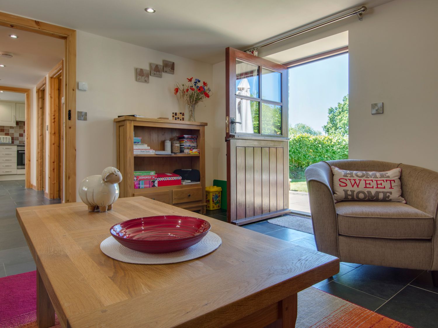 A living room with a wooden coffee table holding a red bowl and a sheep ornament next to an armchair with a Home Sweet Home cushion and an open door leading outside at Orchard Barn in St Giles On The Heath