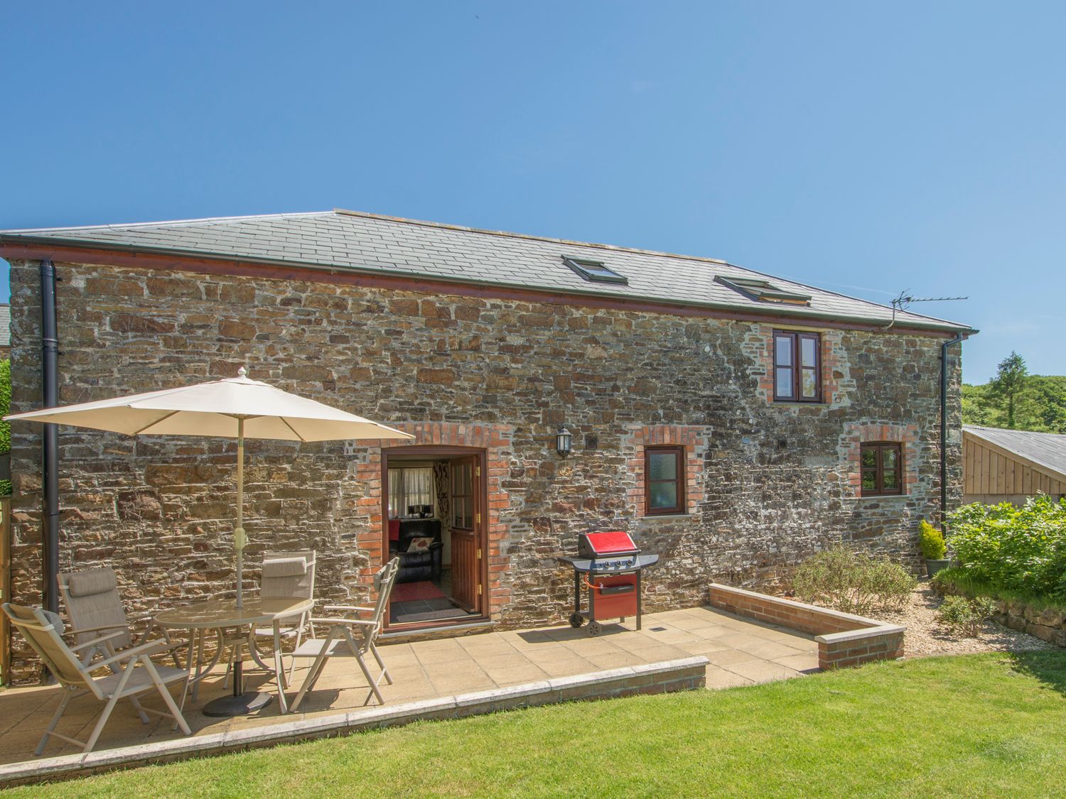 An outdoor patio area with chairs a table an umbrella and a grill next to a stone house at Orchard Barn in St Giles On The Heath