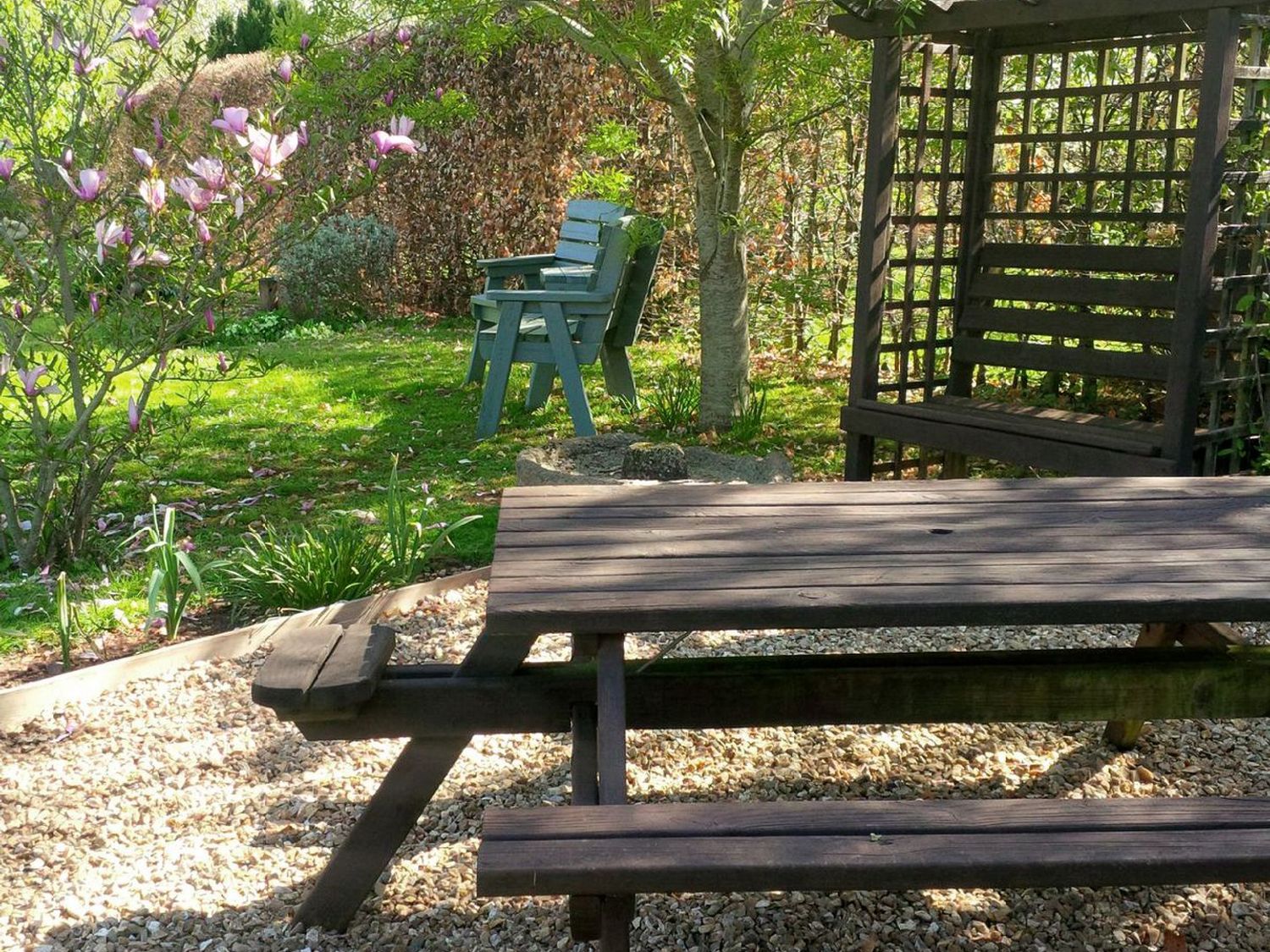 An outdoor garden area with a wooden picnic table bench a wooden garden swing and two green chairs near a tree at The Woodshed in Upton Pyne