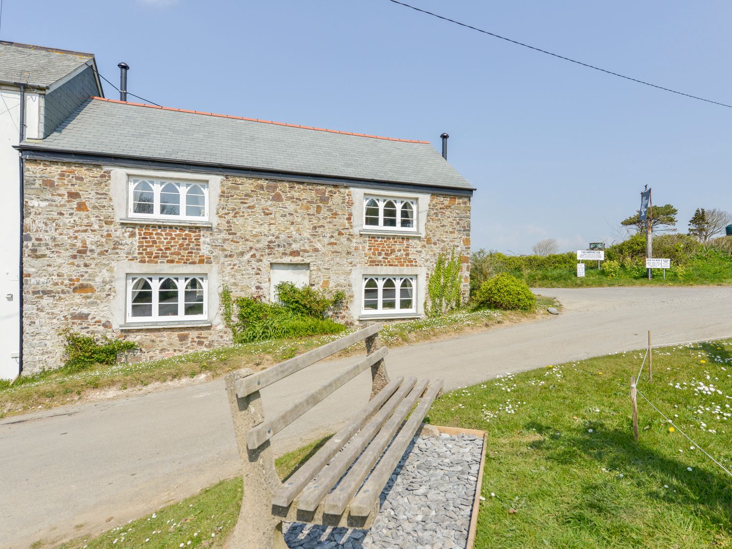 A stone cottage with white framed windows near a gravel road and a wooden bench on grass at Chapel Cottage in Welcombe