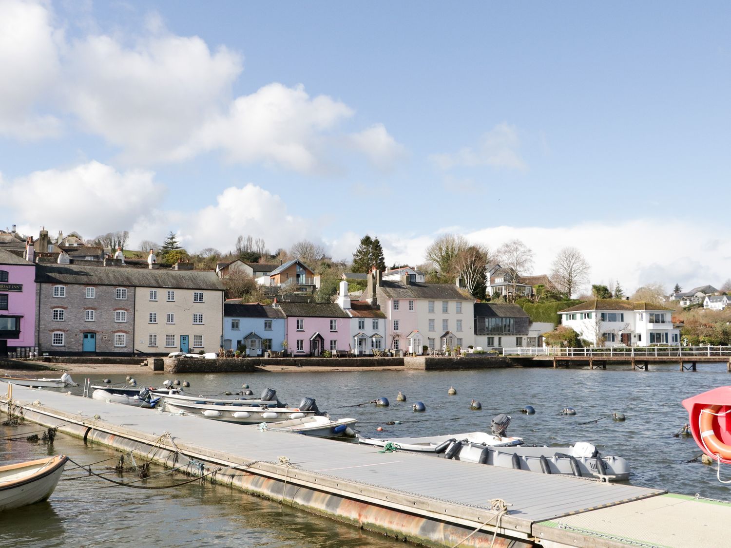 A waterfront with a dock and several small boats moored and houses along the shoreline at Dove Cottage in Dittisham