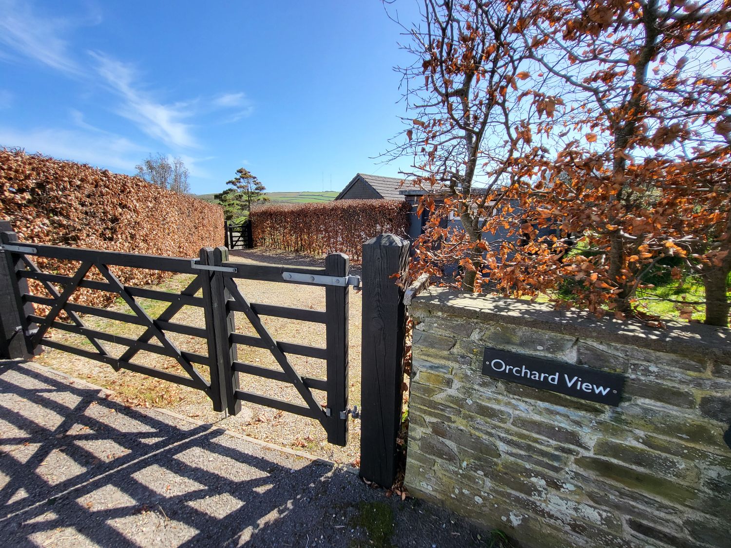 A black wooden gate at the entrance to a gravel driveway with hedges on both sides at Orchard View in Kellaton near Hallsands