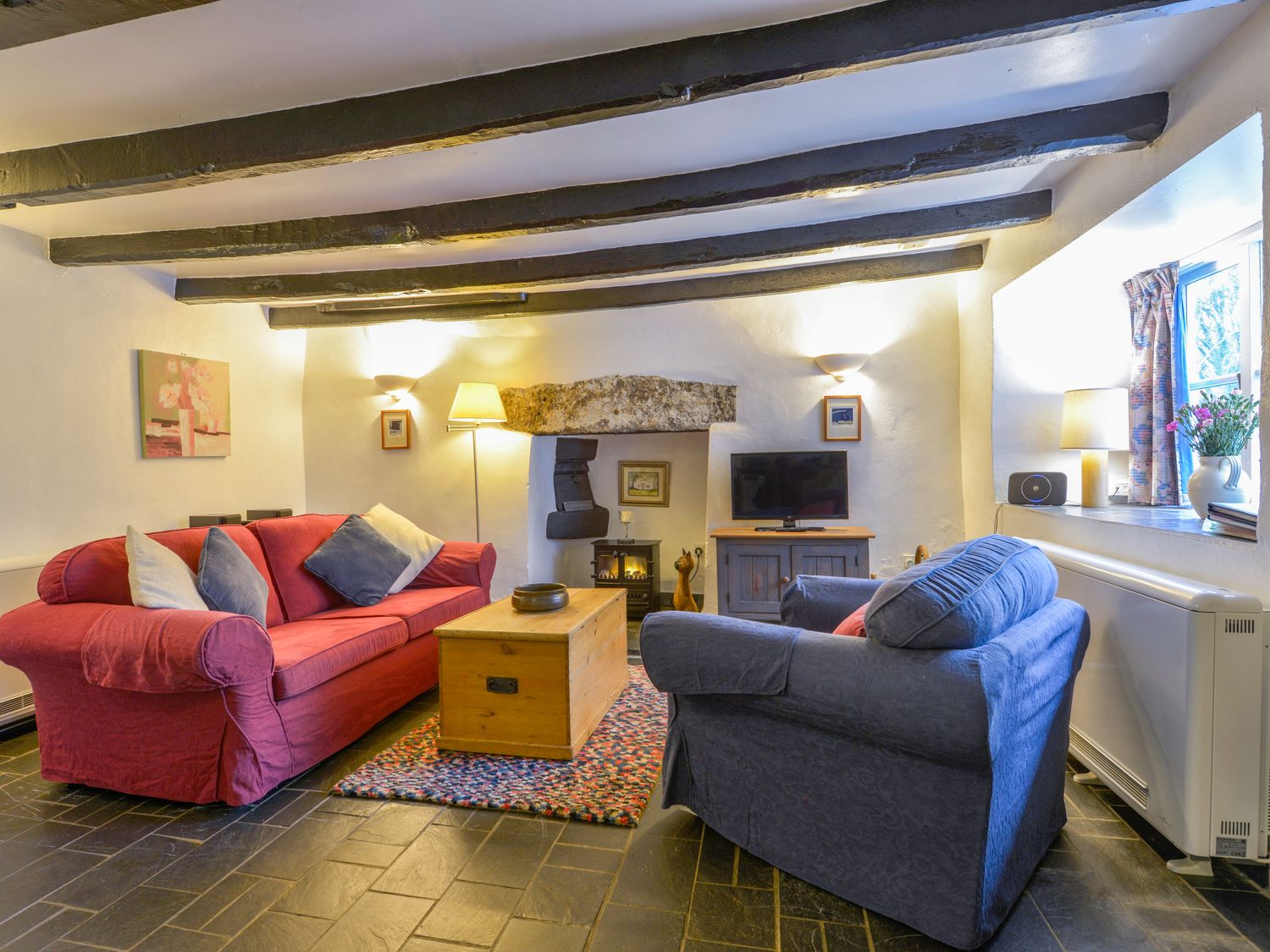A living room with a red sofa and blue armchair a wooden chest table a television and a fireplace at Churchgate Cottage in Blisland