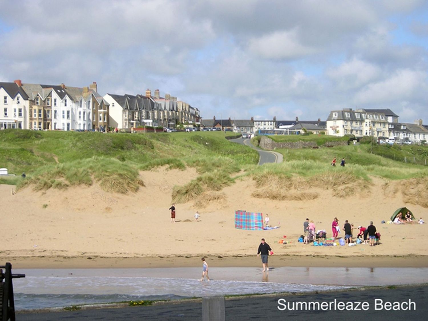 A sandy beach with people near the water and dunes with grass behind houses in the background at Summerleaze Beach