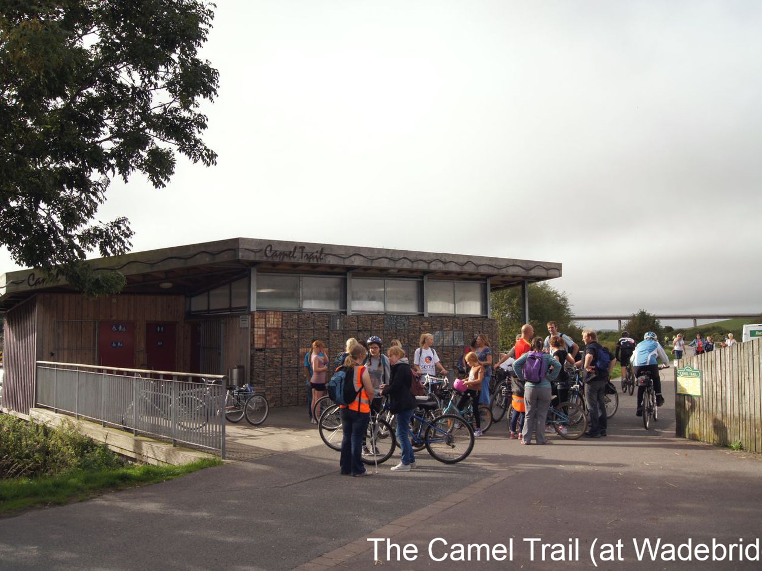 A group of people with bicycles gathered near a small building with signage at The Camel Trail at Wadebridge