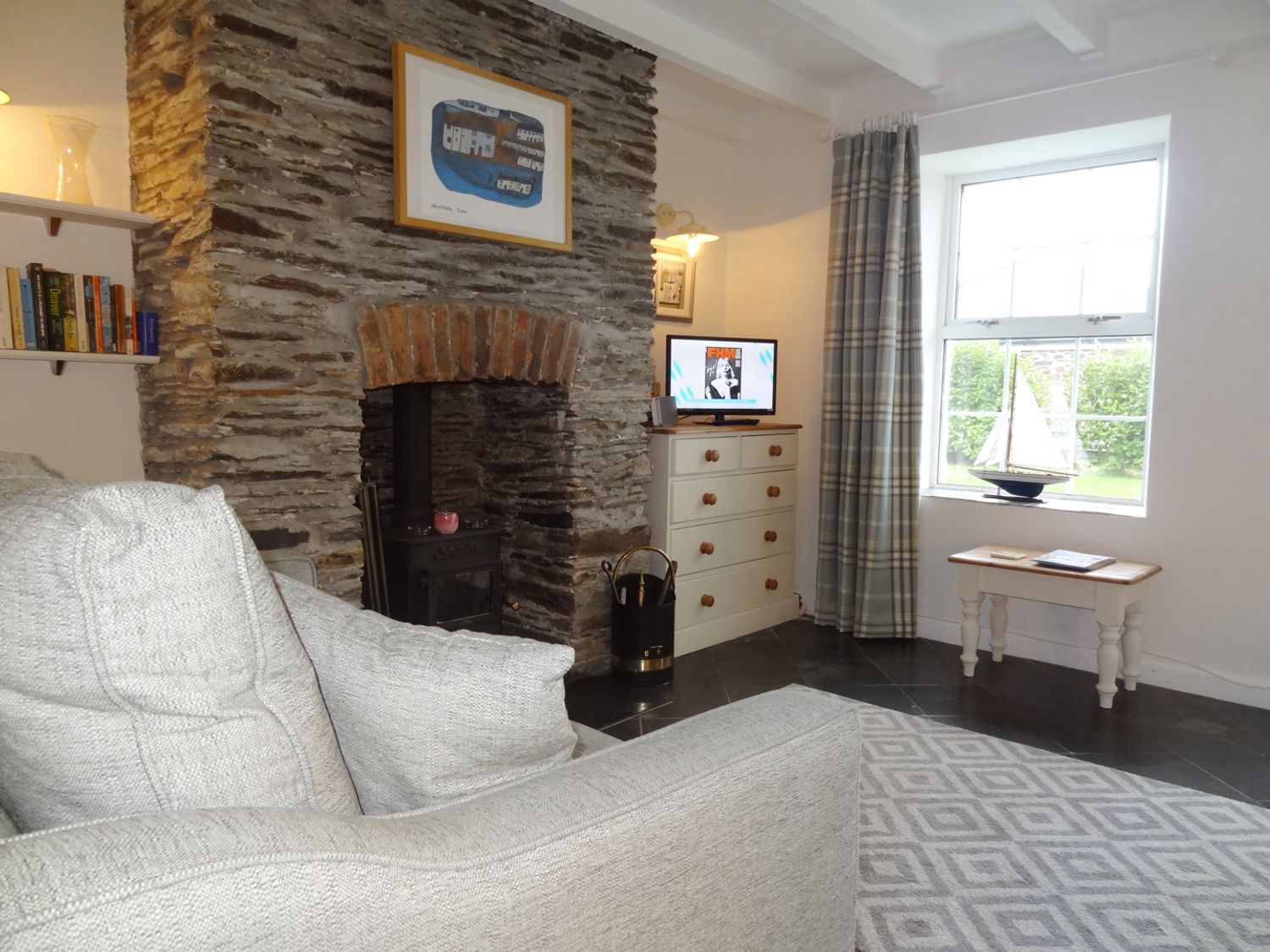 A living room with a stone fireplace a grey sofa a chest of drawers and a window with a model sailboat at Kingfisher Cottage in Edmonton near Wadebridge