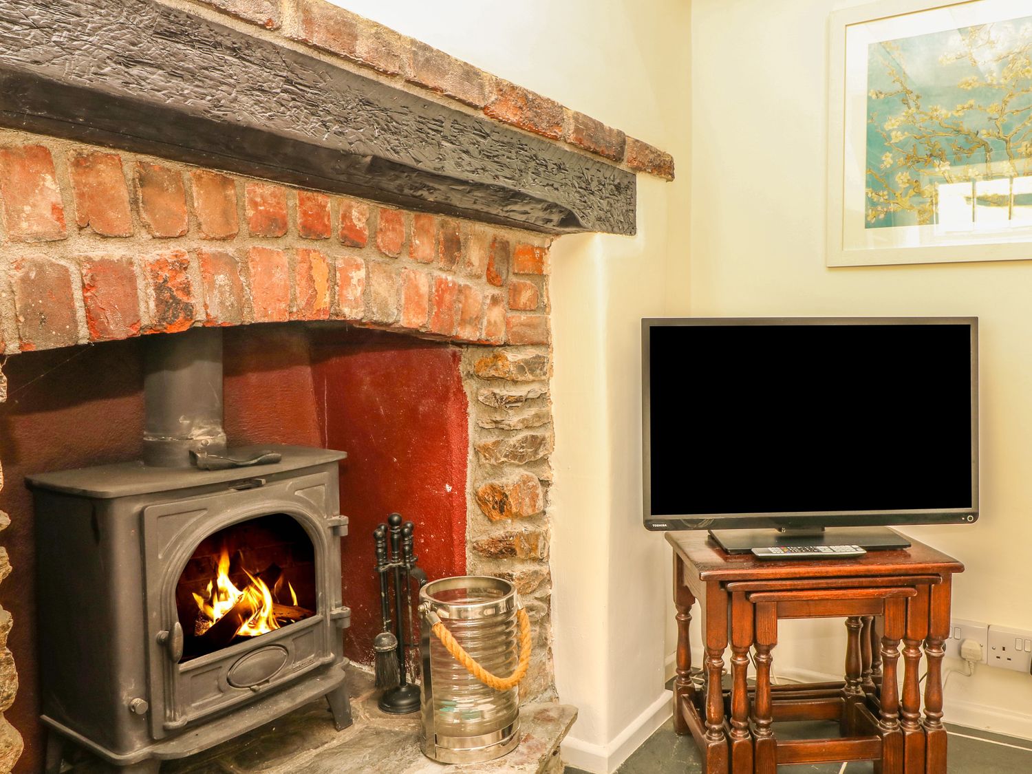 A wood burning stove with fire in a brick fireplace next to a wooden nest of tables with a flat screen tv on top at Cousham Cottage in Kingsand And Cawsand