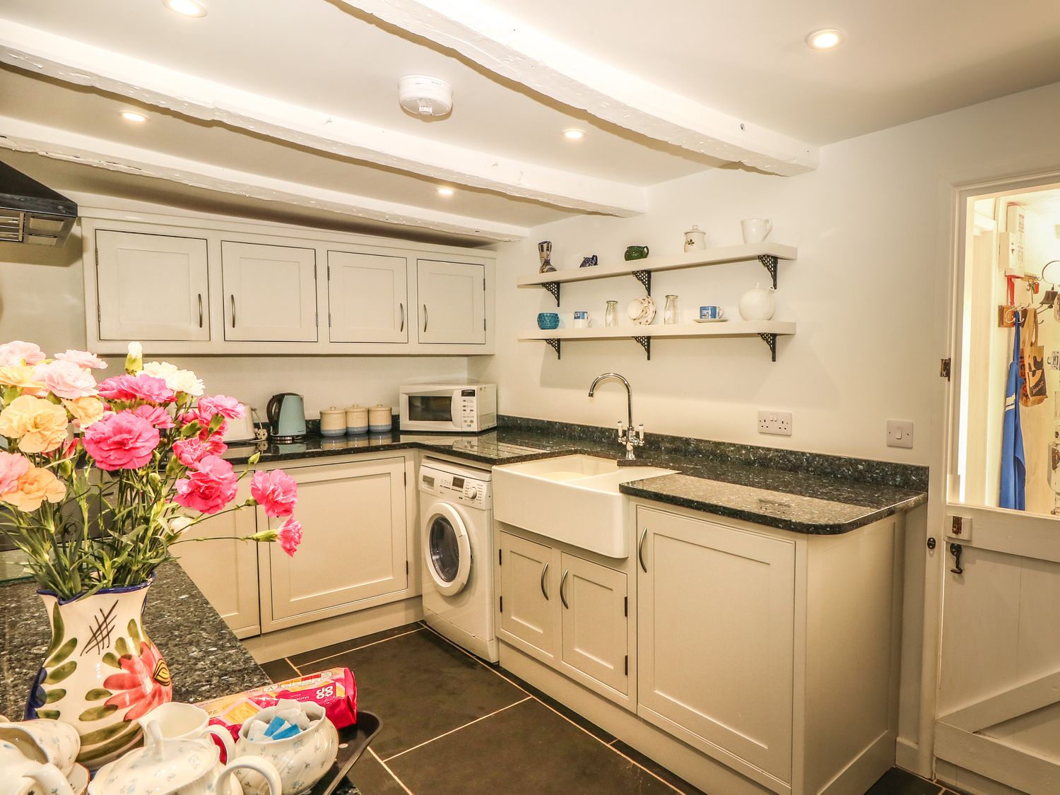 A kitchen with a vase of flowers on the counter washing machine microwave and shelves with cups at Cousham Cottage in Kingsand And Cawsand