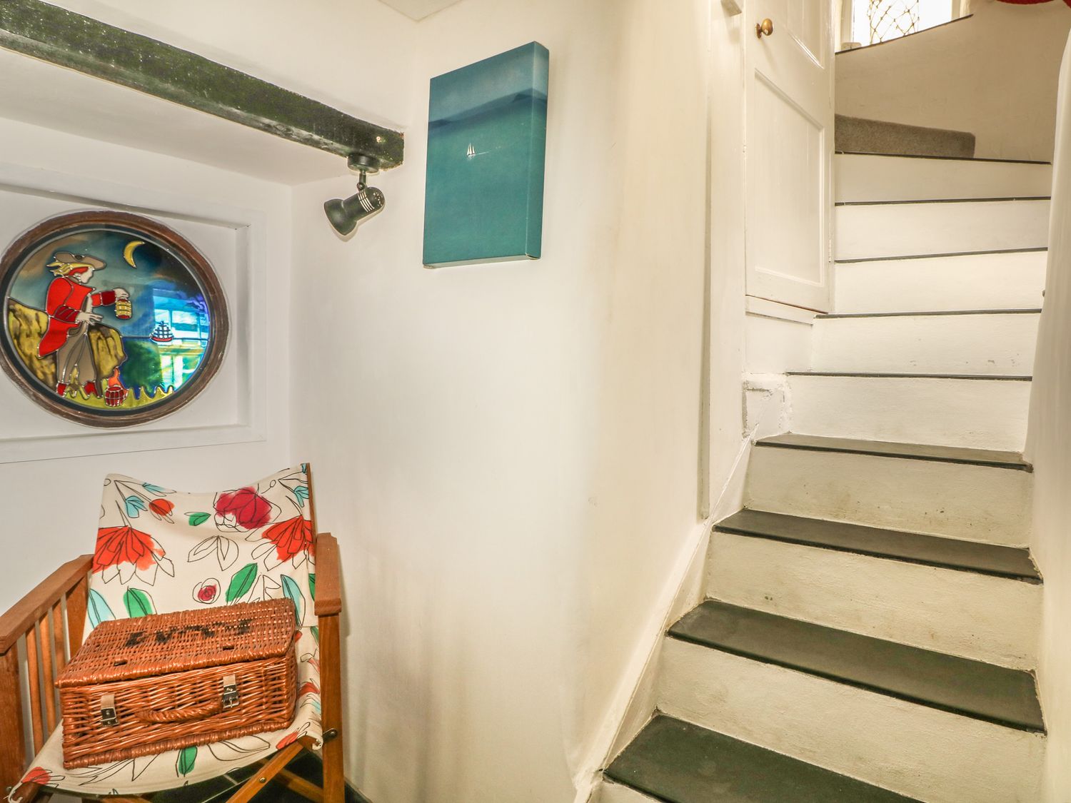 White stairs with a floral chair and a wicker basket below a round stained glass window at Cousham Cottage in Kingsand And Cawsand