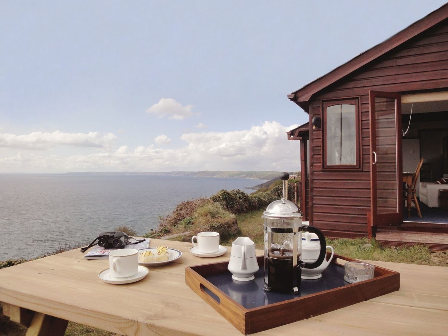 A picnic table with coffee cups, a French press, biscuits, binoculars, and a wooden cabin with an open door by the seaside at Seagulls Nest Whitsand Bay near Millbrook