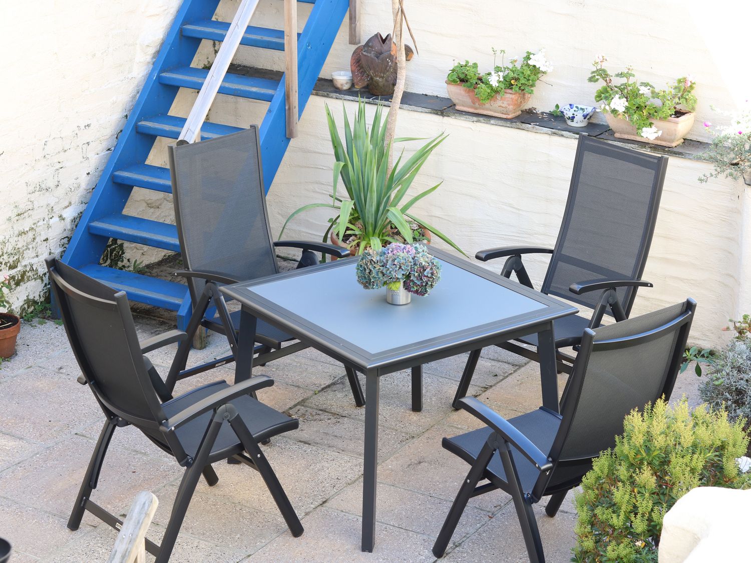 An outdoor patio with a square table and four black chairs near blue stairs and plants at Pier Cottage in St Mawes