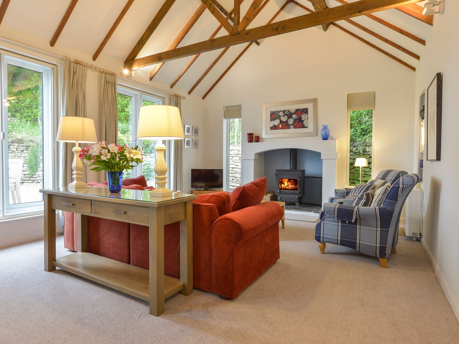 A living room with a wood table with two lamps and flowers a red sofa a blue plaid armchair a fireplace and large windows at Little Barn Cottage in Portloe