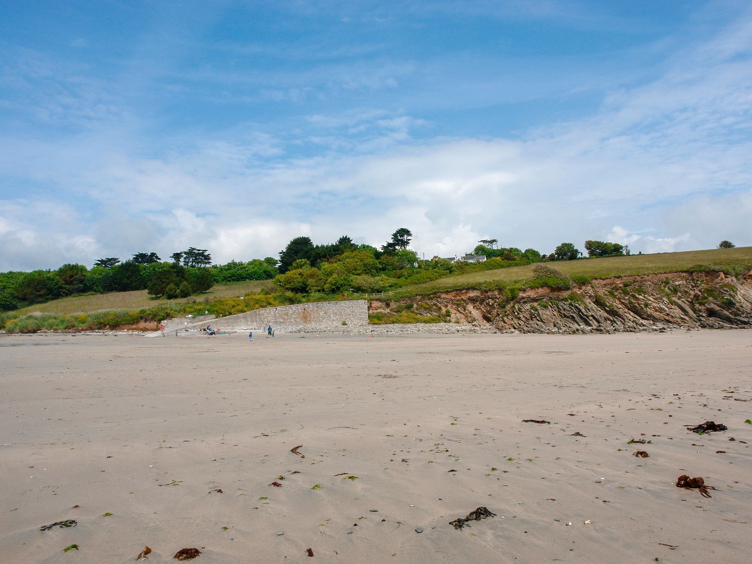 A sandy beach with a stone wall and grassy cliff under a partly cloudy sky at Little Barn Cottage in Portloe
