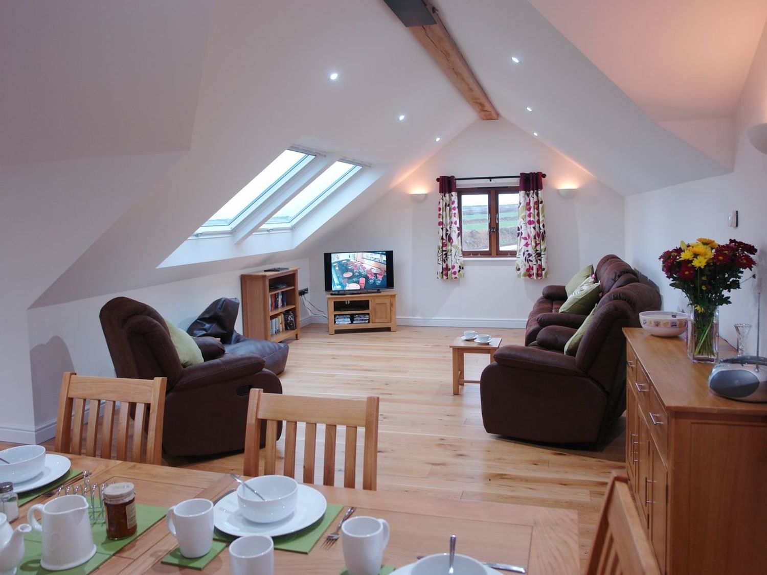 An attic living room with sofas a television dining table set and flowers on a cabinet at The Hayloft in Breage