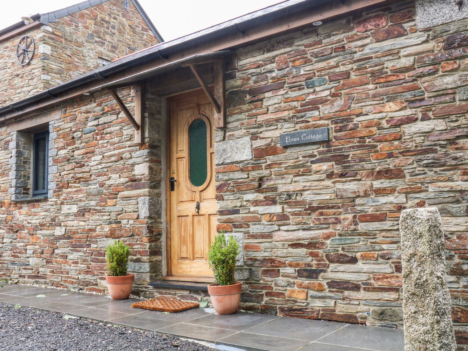 The entrance of a stone cottage with a wooden door two potted plants a doormat and a sign saying Elvan Cottage in Liskeard