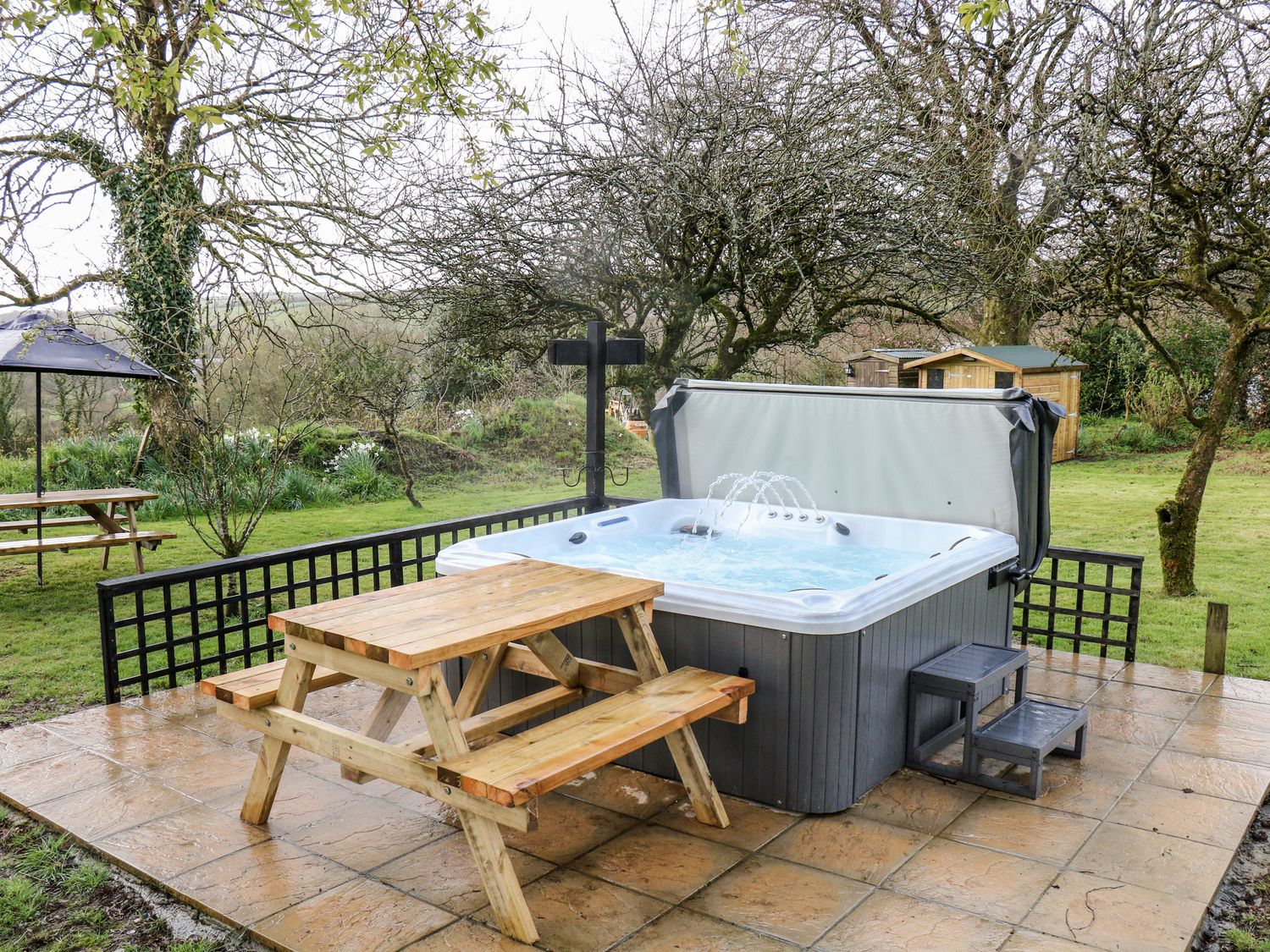 An outdoor patio area with a hot tub, wooden picnic table, and small steps surrounded by grass and trees at Elvan Cottage in Liskeard