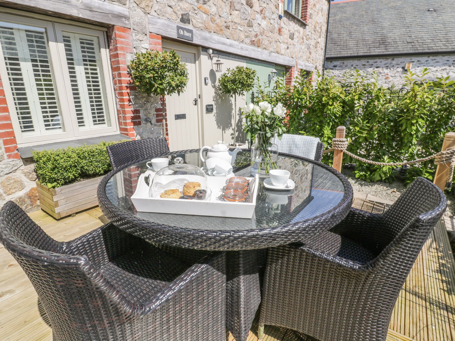 An outdoor patio area with a round glass table and four wicker chairs a tray with teapot scones and cups and a vase with flowers in front of a stone building at Oh Buoy Charlestown