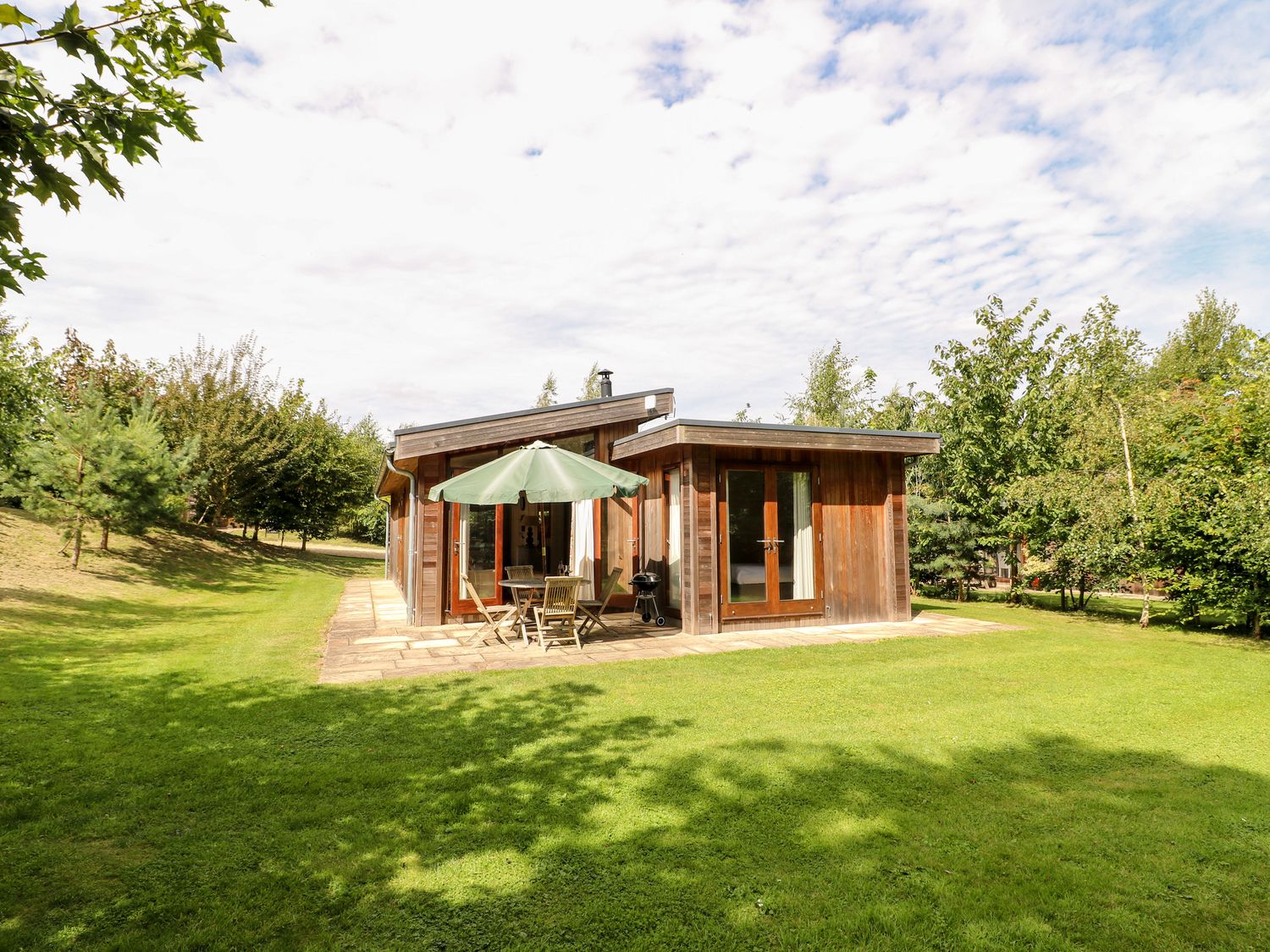 A small wooden house with a patio table and umbrella on a grassy lawn surrounded by trees at Tickencote in Exton Rutland