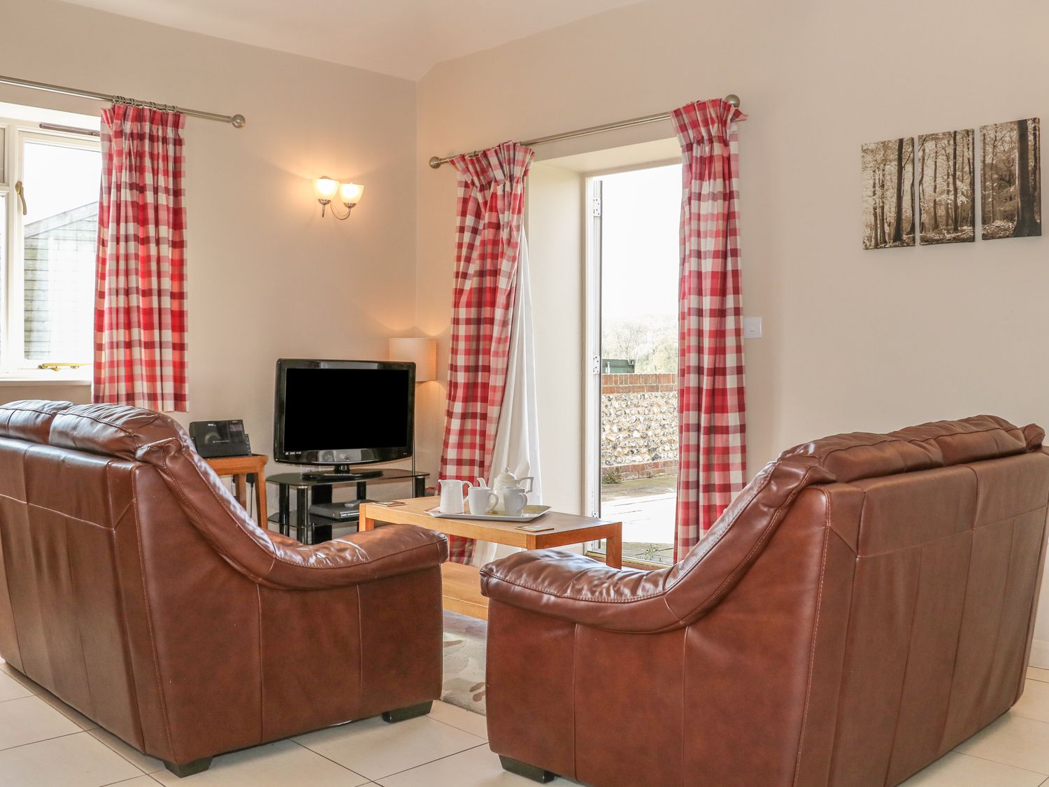 A living room with two brown leather sofas a wooden coffee table a television and red and white check curtains at The Old Dairy in Angmering