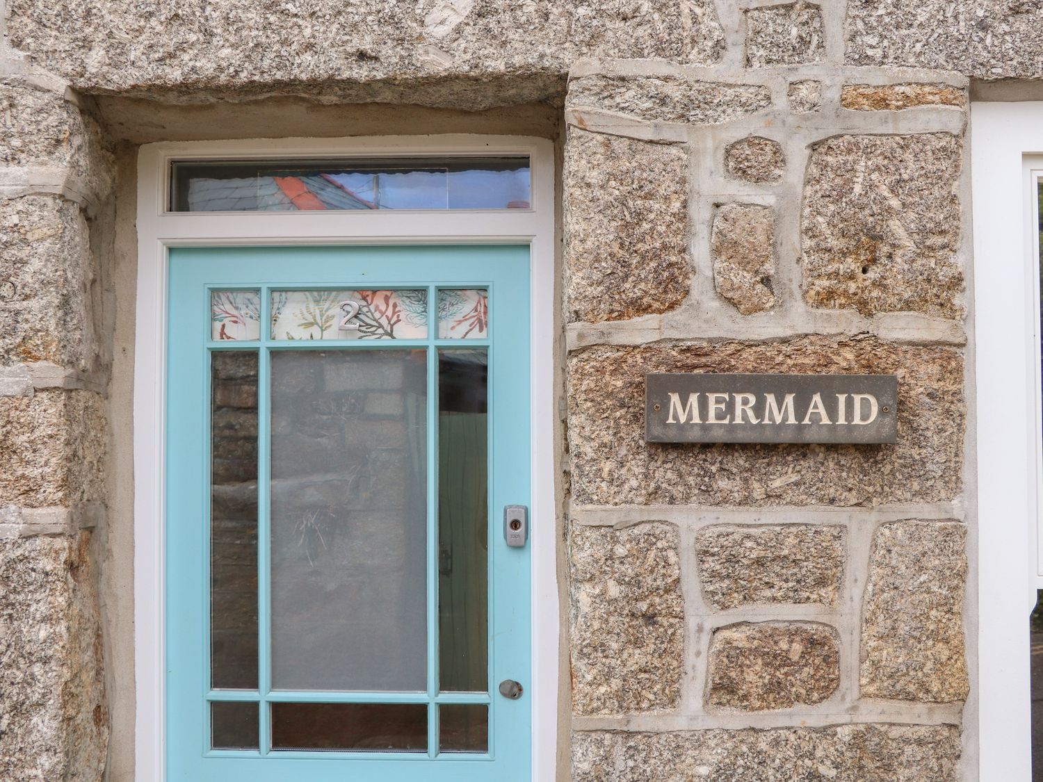 A stone exterior wall with a blue door and a sign that reads Mermaid at Mermaid Cottage in Mousehole
