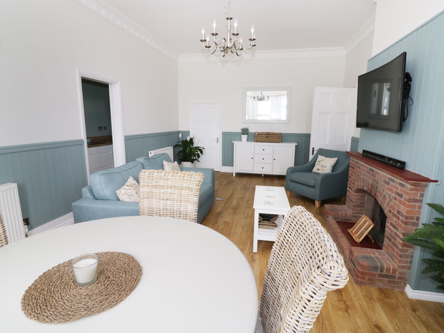 A living room with blue couches a brick fireplace a wall-mounted TV and a dining table with wicker chairs at Flat 1 Cambridge Court Frinton-on-Sea near Walton-on-the-Naze