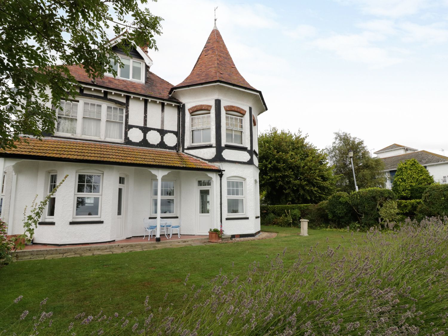 The exterior of a white two-story house with a pointed turret and a garden with green grass and bushes at Flat 1 Cambridge Court in Frinton-on-Sea near Walton-on-the-Naze