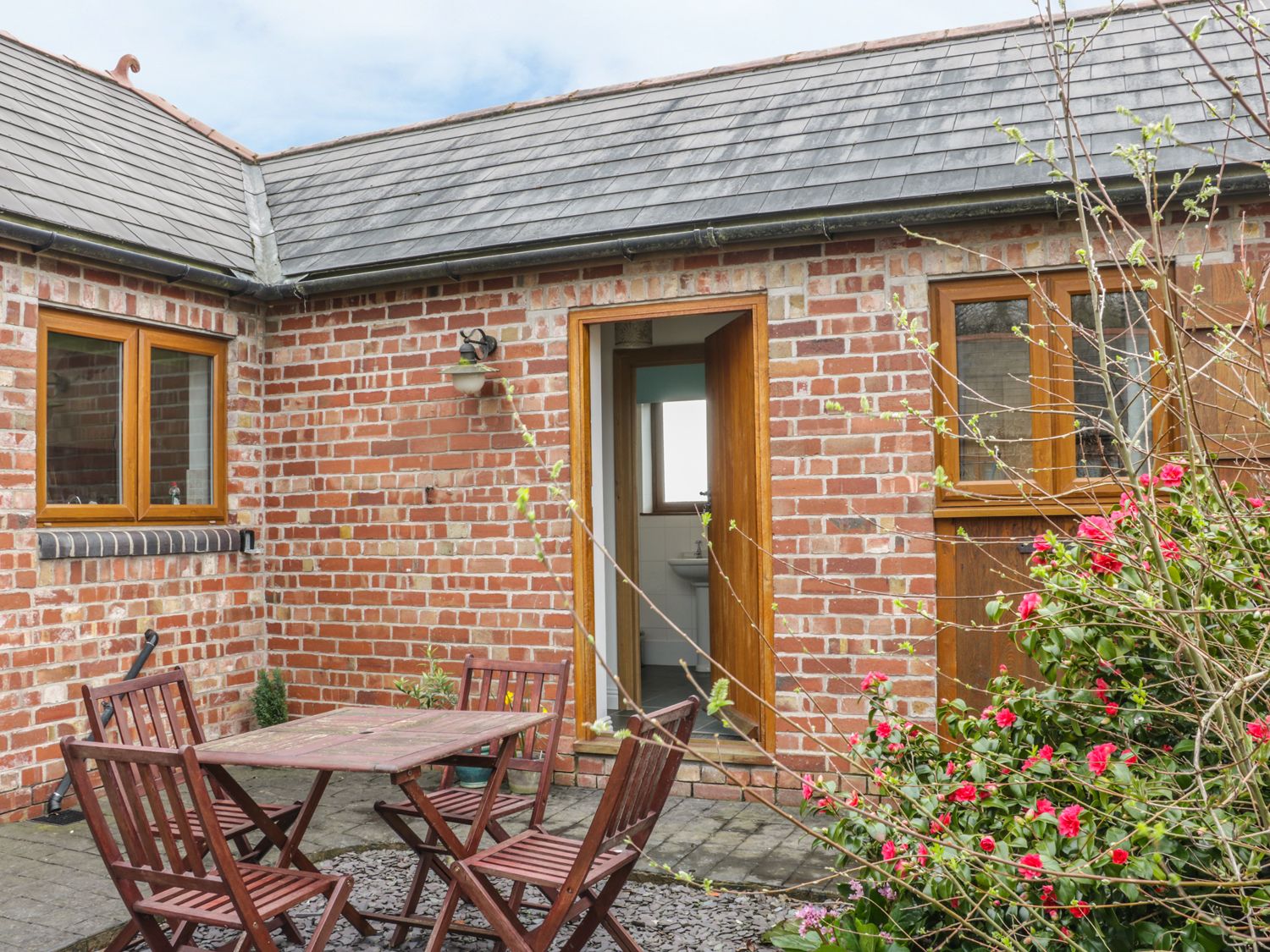 An outdoor patio with wooden table and chairs next to a brick building with two windows and an open door at Acorn Cottage 2 Burlton near Ellesmere