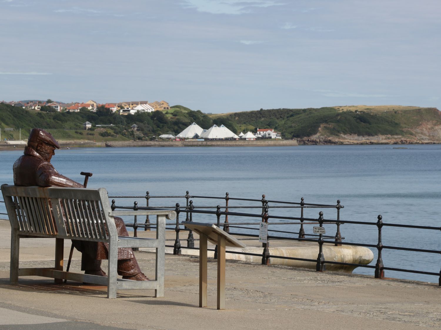 A statue of a seated person on a bench by the waterfront with hills and white tents in the background at Hayloft in Staintondale