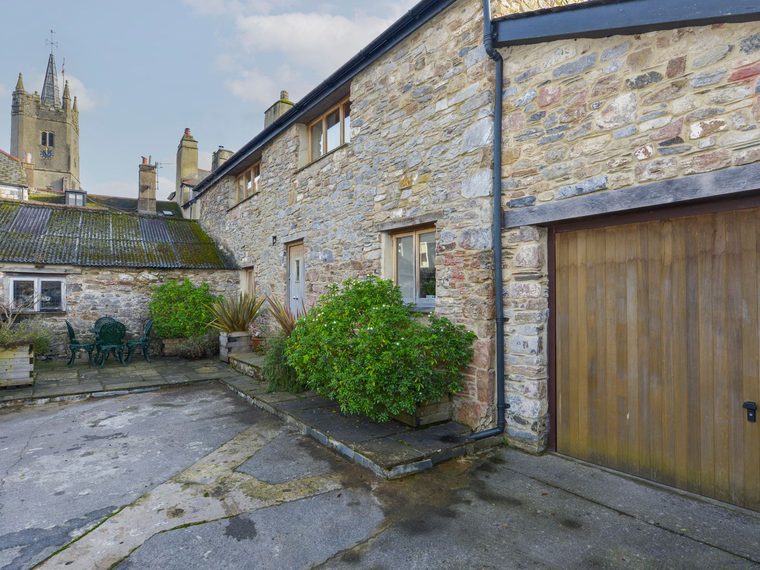 An outdoor patio area with stone walls wooden garage door table with chairs and potted plants at Old Armoury Barn in Ashburton