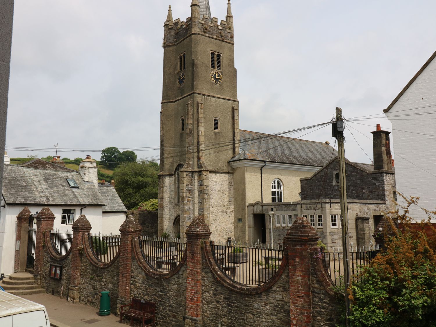 A stone church with a clock tower behind a brick and stone wall with iron fencing in a village at The Armourers Forge in Ashburton