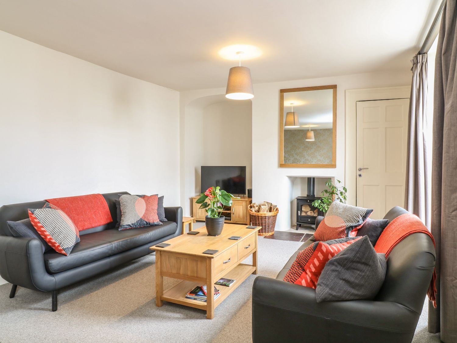 A living room with two black sofas with red and gray cushions a wooden coffee table with a plant a television a basket with firewood and a fireplace at The Armourers Forge in Ashburton
