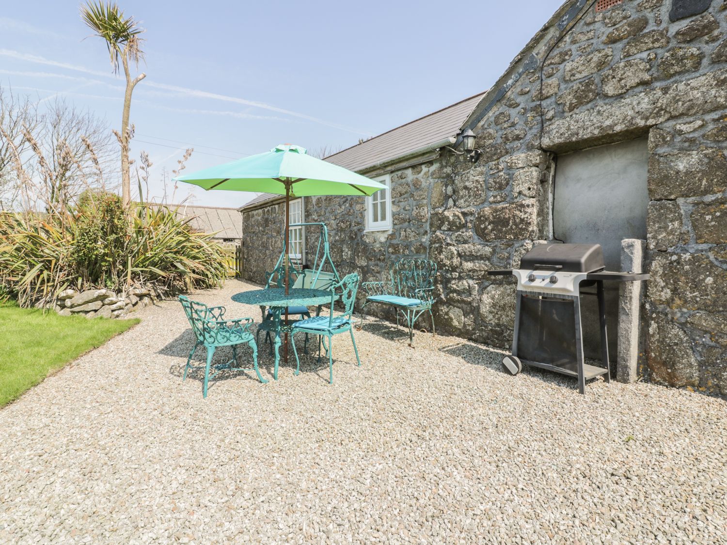An outdoor patio area with turquoise metal furniture including chairs a bench and a table with an umbrella and a black barbecue grill next to a stone building at Anjarden Byre in Newlyn