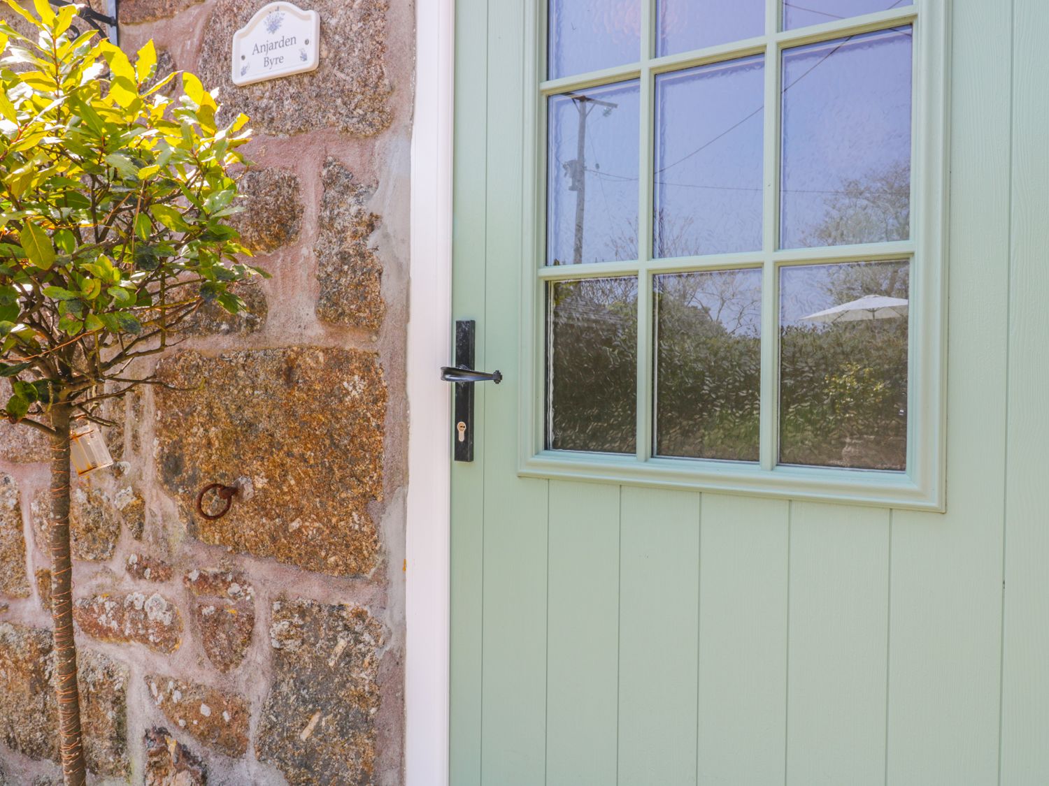 A green door with glass panels next to a stone wall with a small tree and a sign reading Anjarden Byre in Newlyn