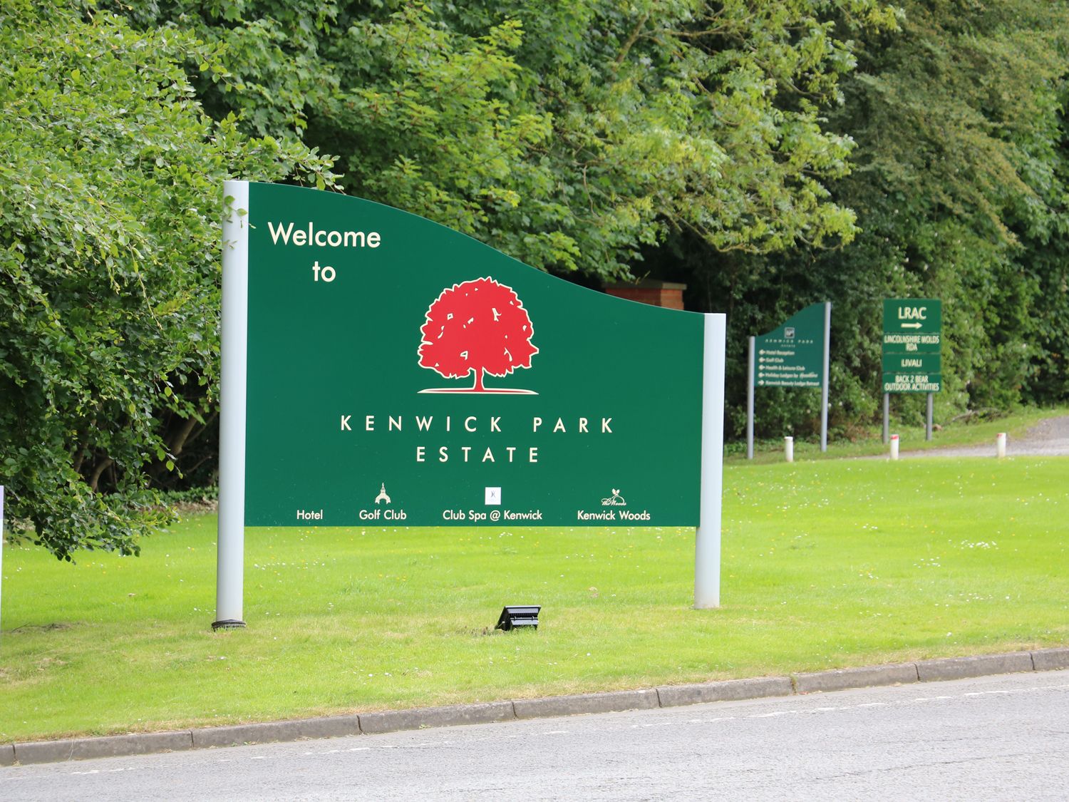 A green welcome sign for Kenwick Park Estate on grass by a road with trees in the background