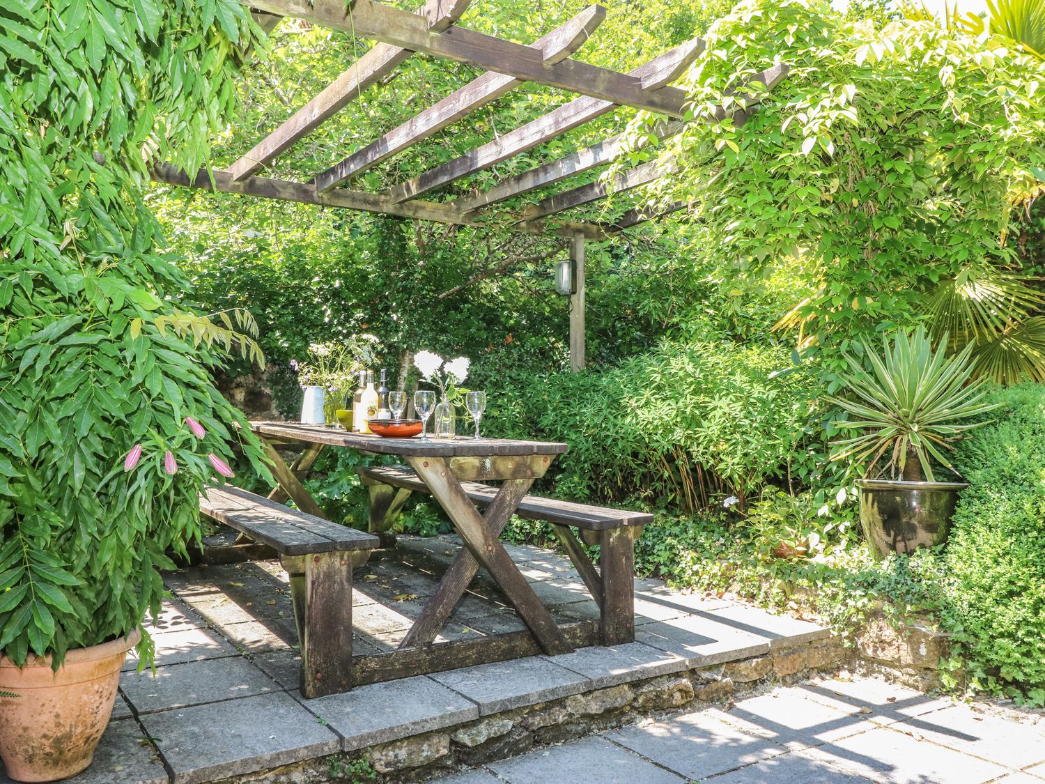 An outdoor garden area with a wooden picnic table and benches under a wooden pergola surrounded by green plants and foliage at The Cider Barn in Loddiswell
