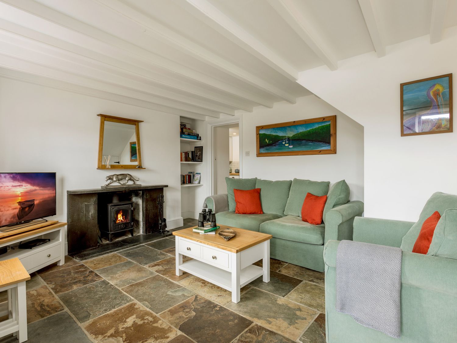 A living room with green sofas red cushions a wood burning stove a coffee table and paintings at Fal River Cottage in Malpas Cornwall