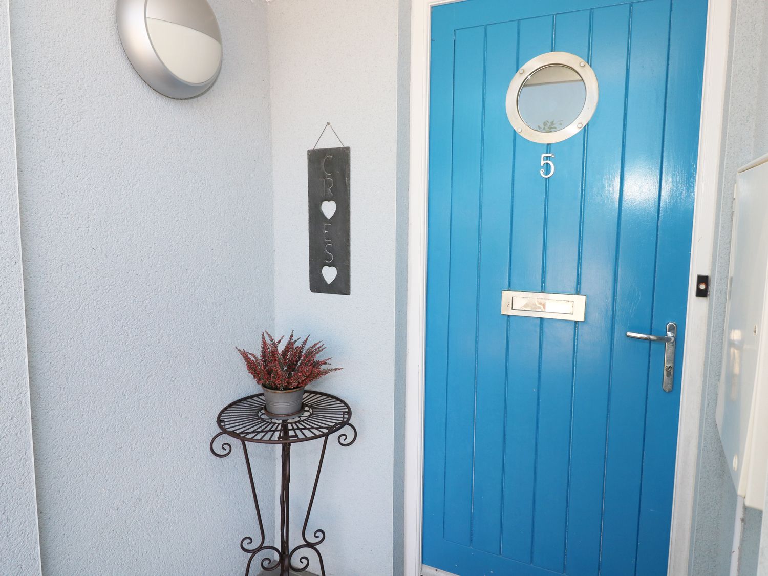 A blue front door with a round window and number 5 next to a metal table with a plant and a welcome sign at 5 Chandlers Yard in Burry Port