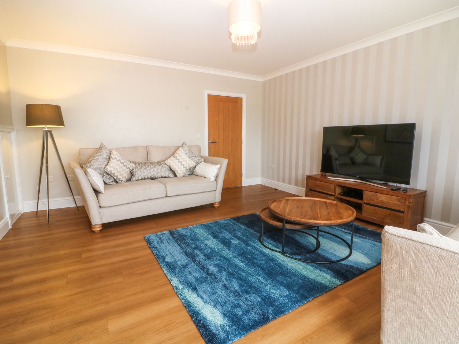 A living room with beige sofas a blue rug wooden coffee tables a TV on a wooden stand and a floor lamp at The Moorings in Y Felinheli