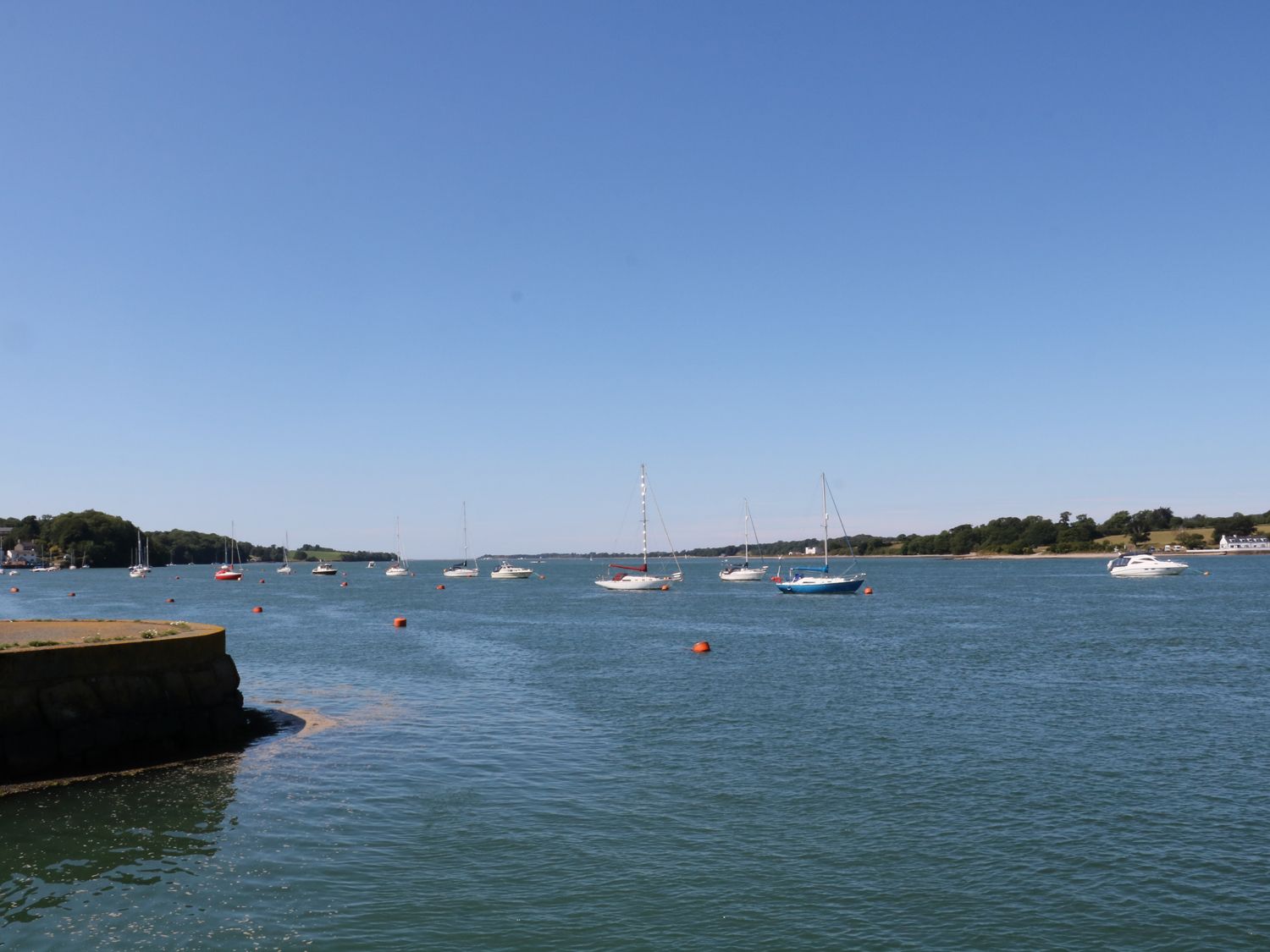 A body of water with sailboats and motorboats anchored near the shore with trees and buildings in the background at The Moorings in Y Felinheli