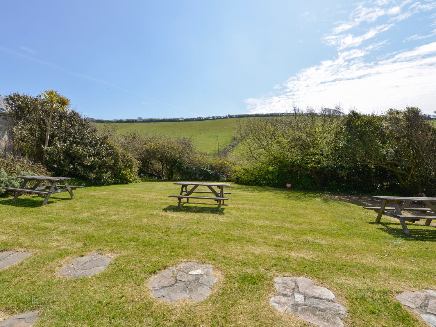 A garden with picnic tables and stone paths surrounded by bushes and fields at 2 Brook Cottages in Hope Cove
